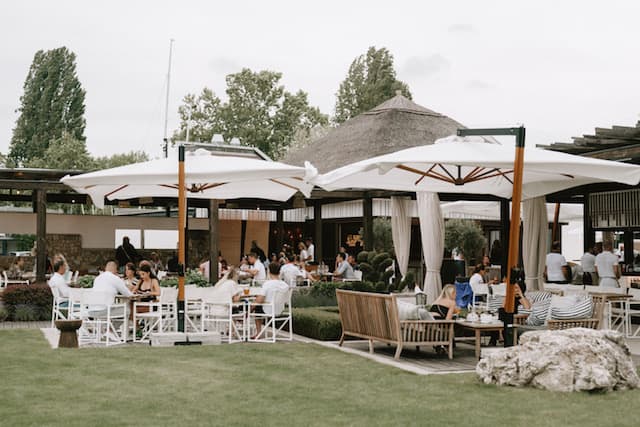 Eine belebte Restaurantterrasse mit weißen Sonnenschirmen, Gästen an Tischen und einem Gebäude mit Reetdach.