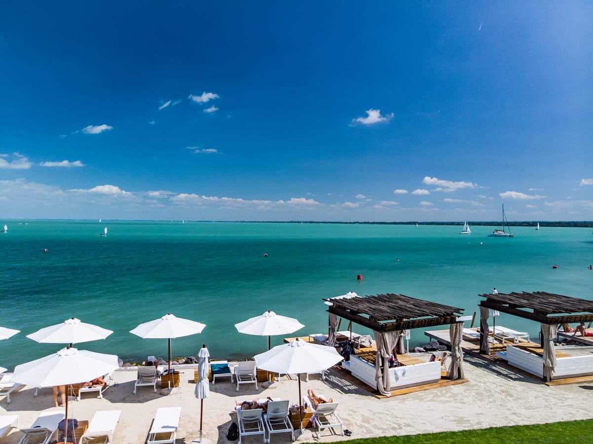 Weiße Sonnenschirme und Holz-Cabanas an einem Sandstrand vor türkisblauem Wasser mit fernen Segelbooten.