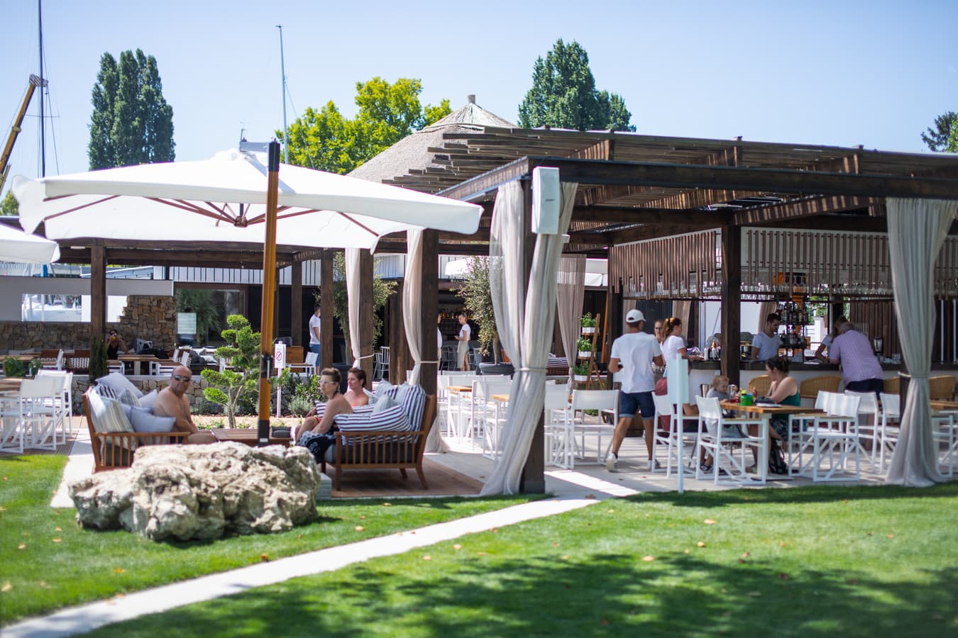 Sonniger Außenbereich eines Strandclubs mit weißen Sonnenschirmen, Holzpavillons und Gästen auf einer gepflegten Rasenfläche.