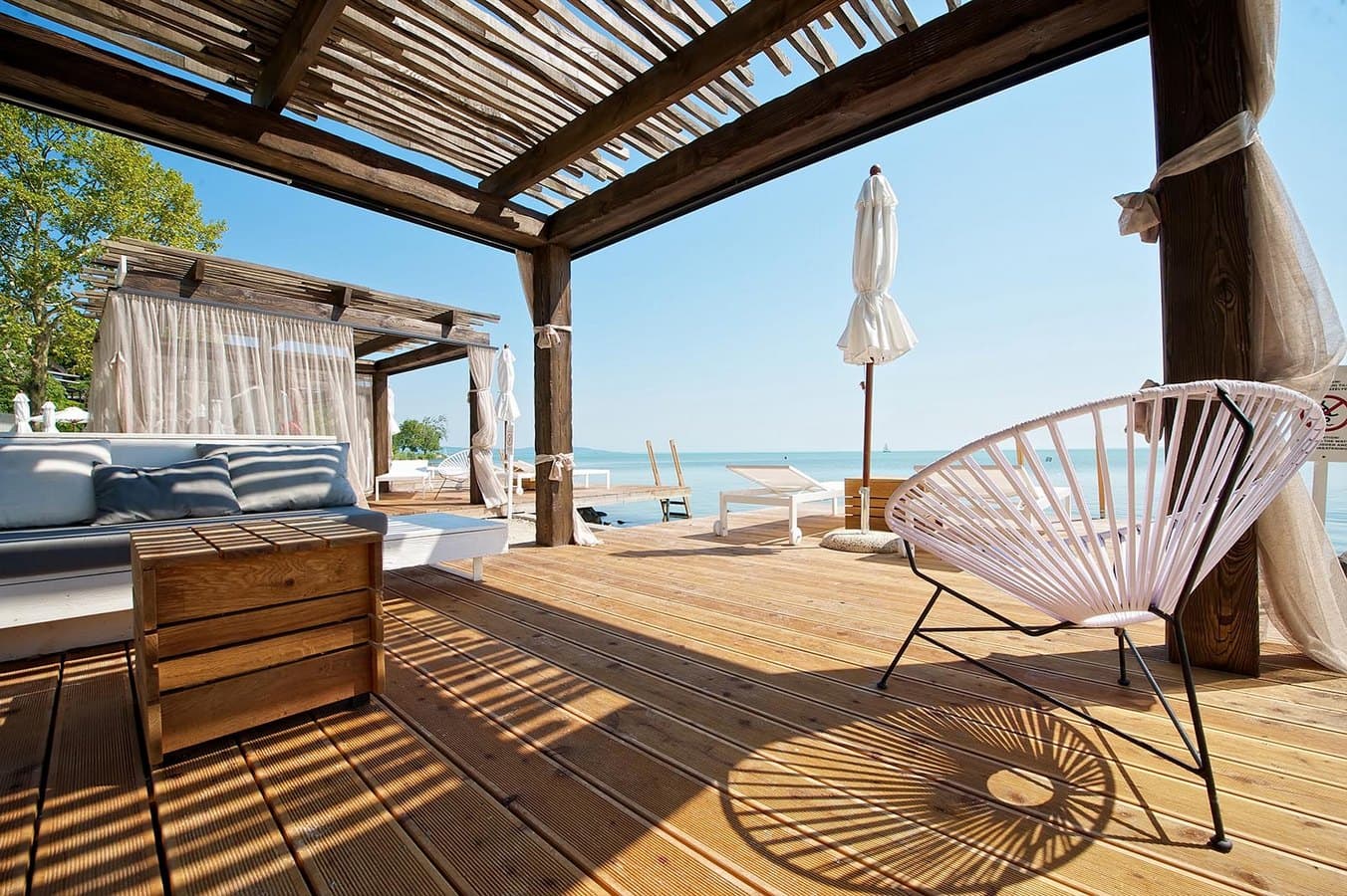 Luxuriöse Holzterrasse am See mit weißen Loungemöbeln, einer Pergola und Blick auf das ruhige Wasser.