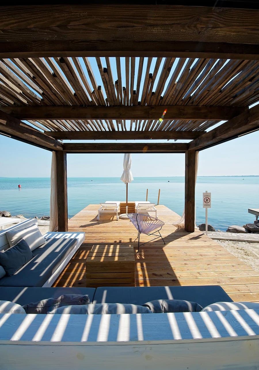 Schattiger Holzpavillon mit Loungemöbeln auf einem Steg mit Blick auf das ruhige, blaue Meer.