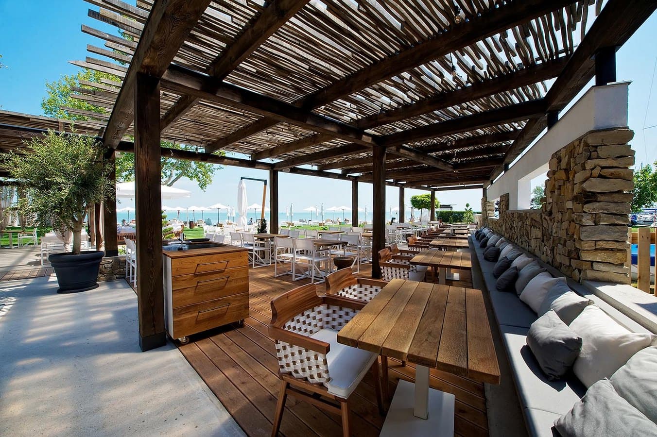 Rustikale Restaurantterrasse unter einer Holzpergola mit Blick auf einen sonnigen Strand mit weißen Sonnenschirmen.