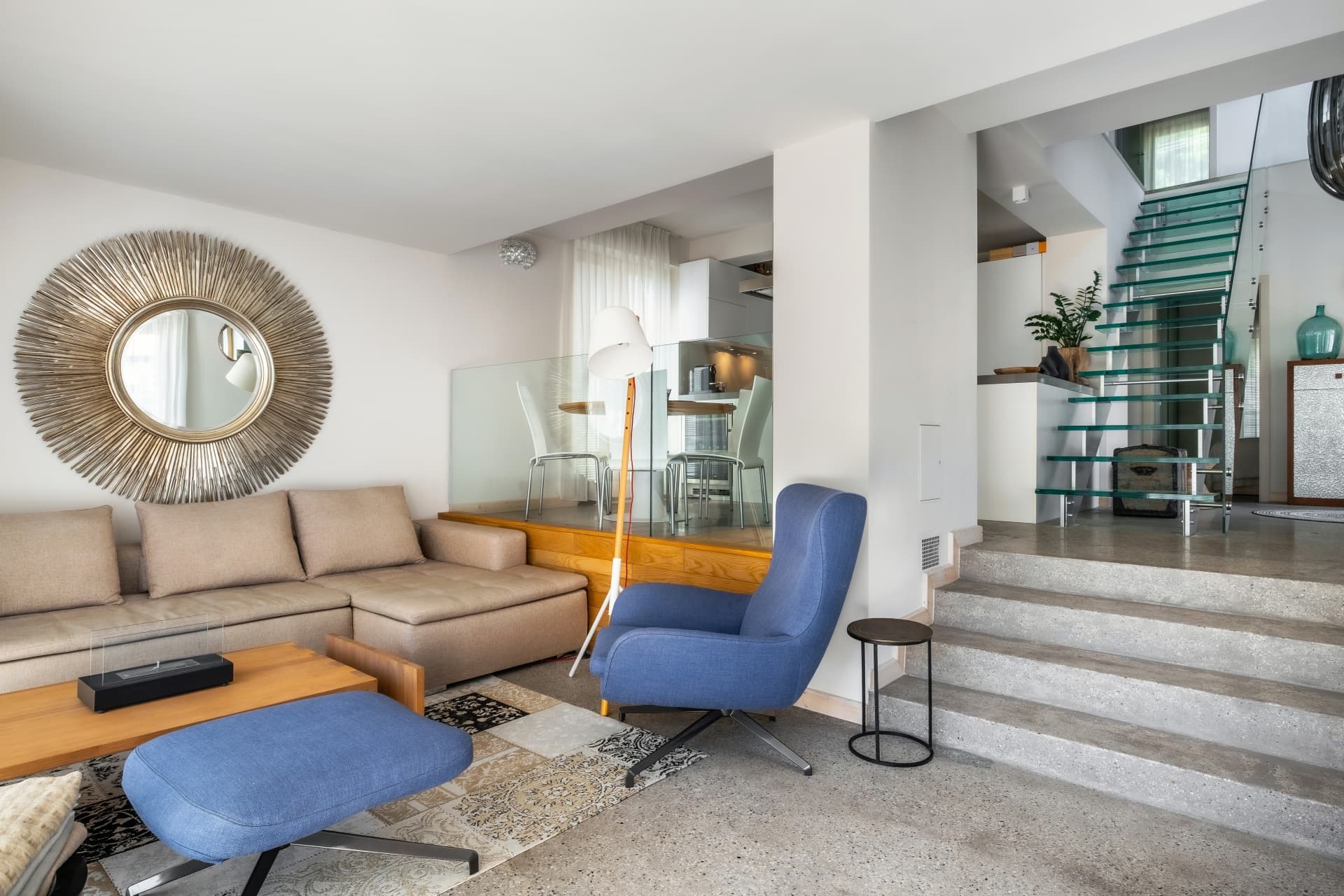 Modernes, offenes Wohnzimmer mit beigem Sofa, blauem Sessel, Sonnenspiegel und Treppen auf mehreren Ebenen.