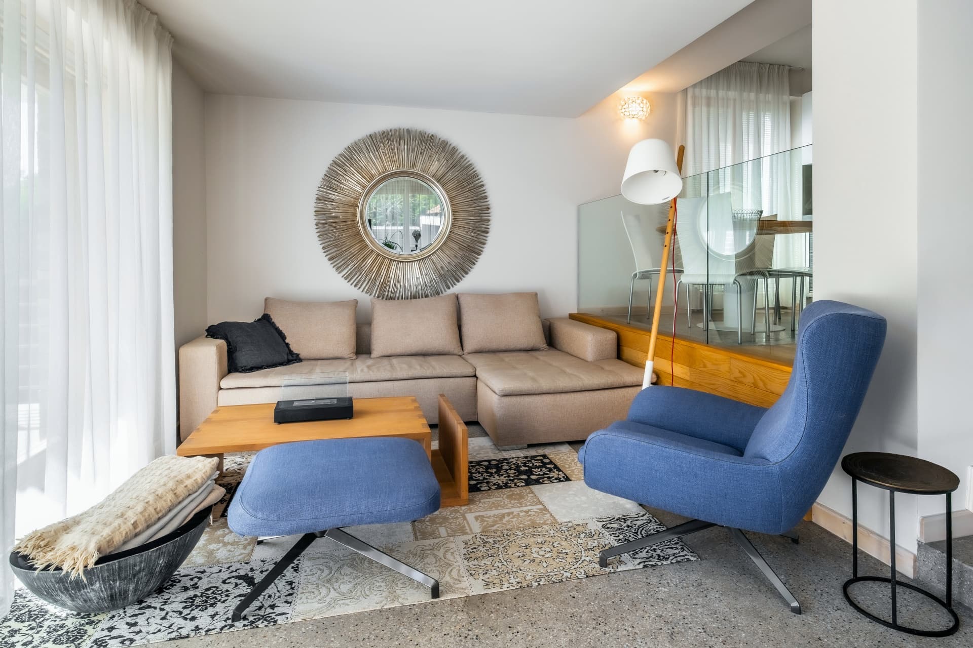 Helles Wohnzimmer mit beigem Ecksofa, blauem Sessel, großem Sonnenspiegel und einem gemusterten Teppich.