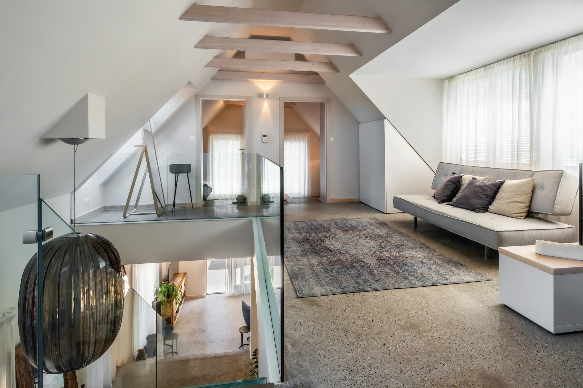 Modernes Dachgeschoss mit grauem Sofa, Holzbalken, gemustertem Teppich und Glasgeländer über einer offenen Treppe.