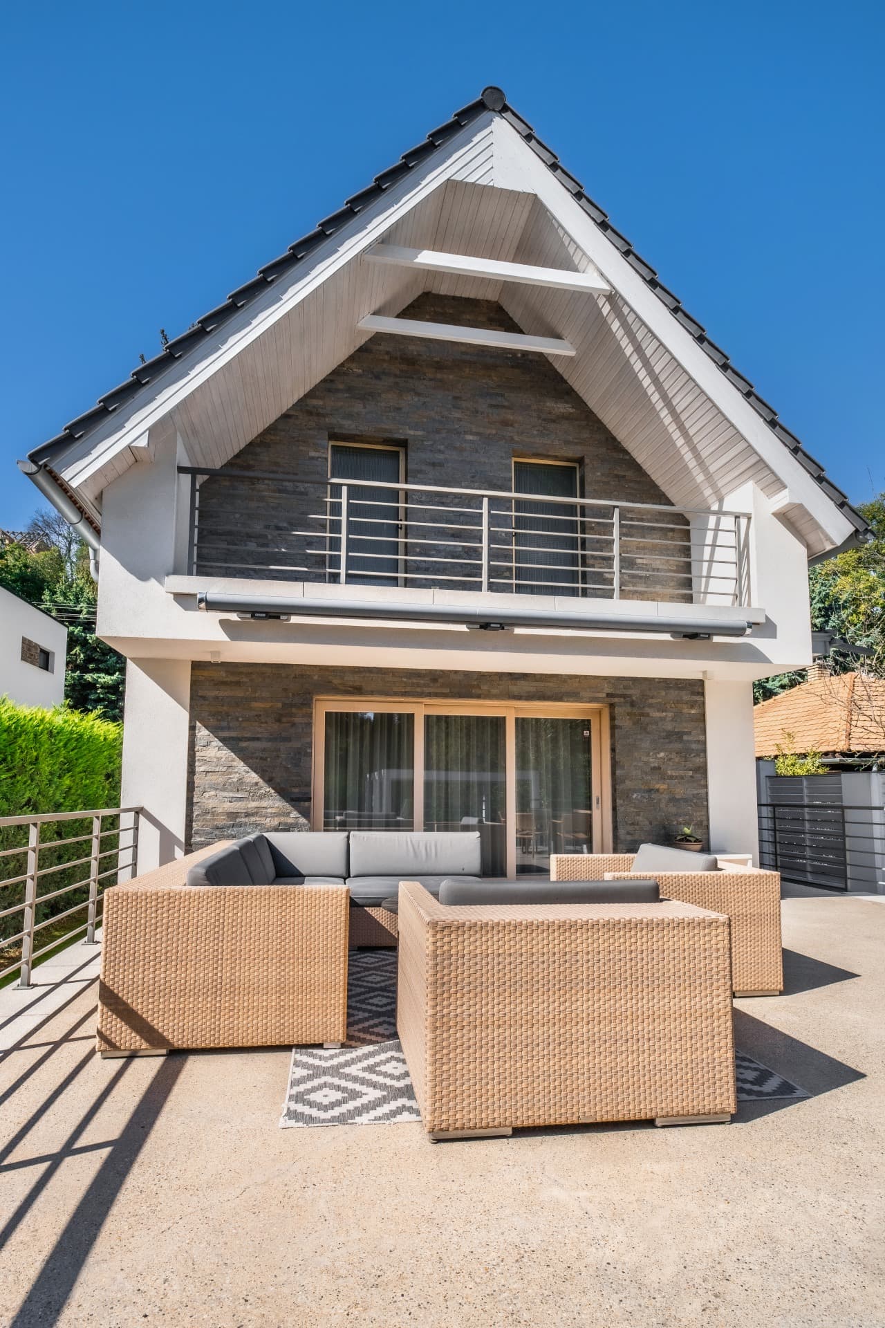 Modernes Haus mit Steinfassade, Balkon und beigen Loungemöbeln auf einer sonnigen Terrasse bei blauem Himmel.