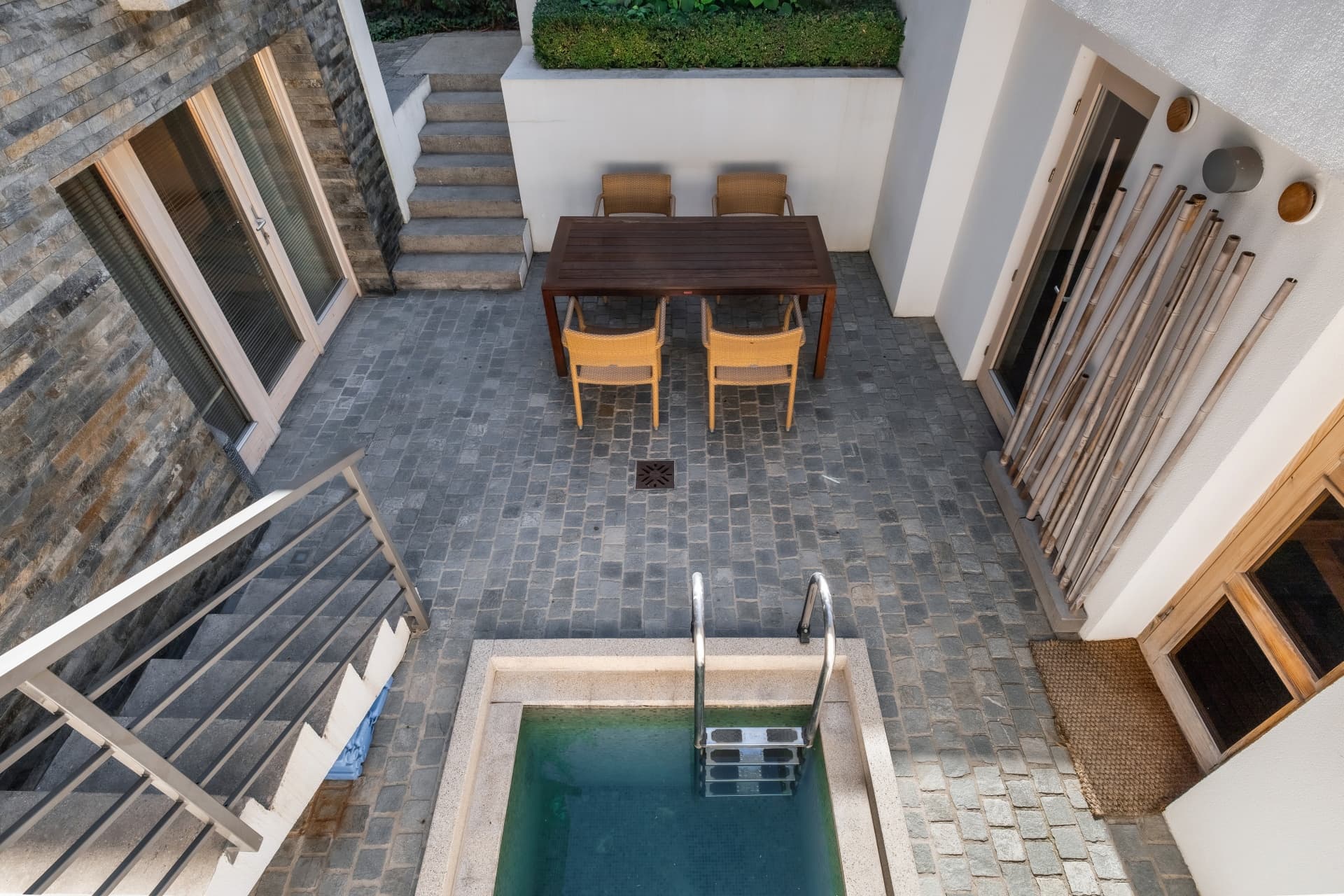 Moderner gepflasterter Innenhof mit kleinem Pool, Holztisch, Korbstühlen und einer Treppe mit Metallgeländer.
