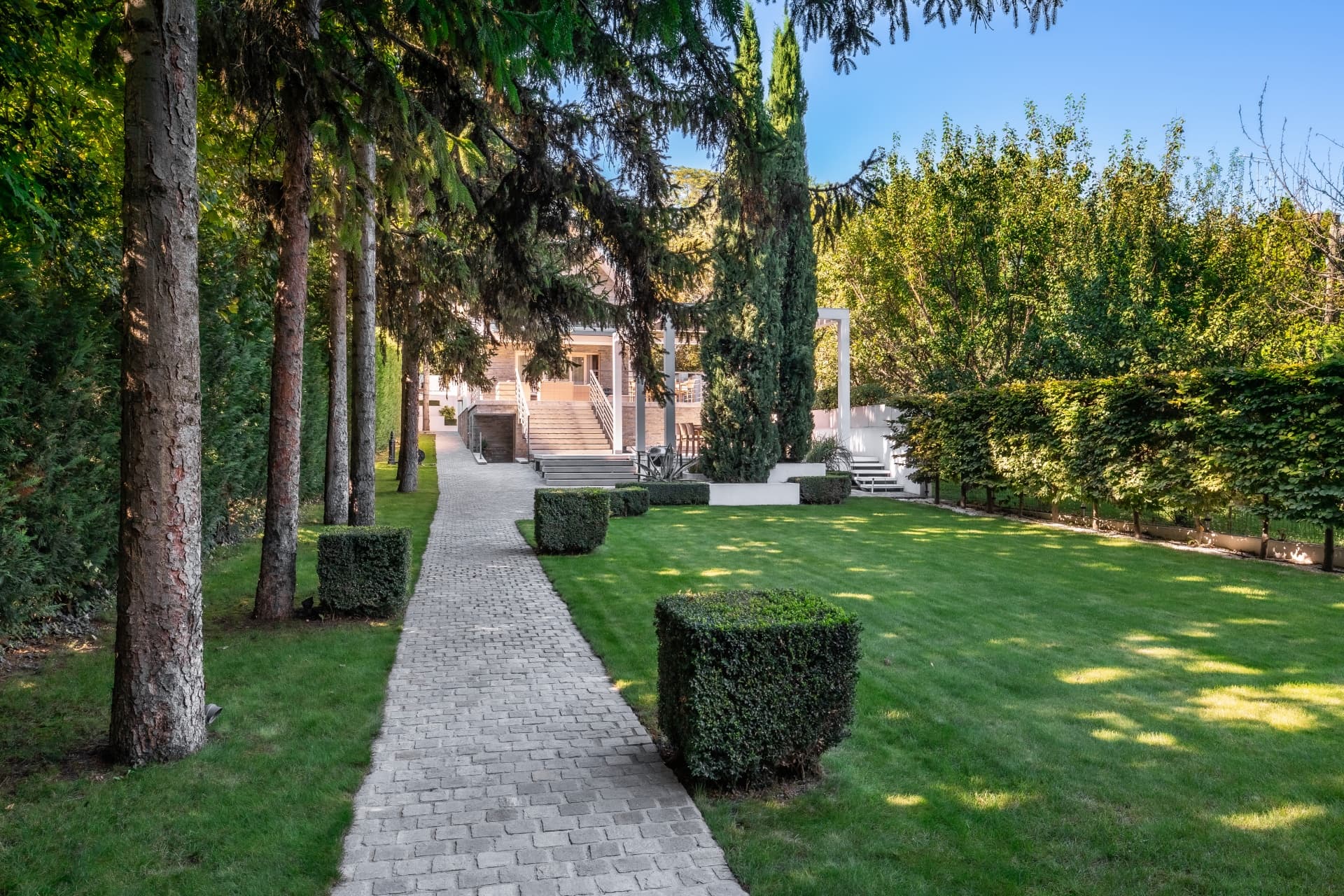 Gepflasterter Weg durch einen gepflegten Garten mit grünem Rasen, quadratischen Büschen und hohen Nadelbäumen.