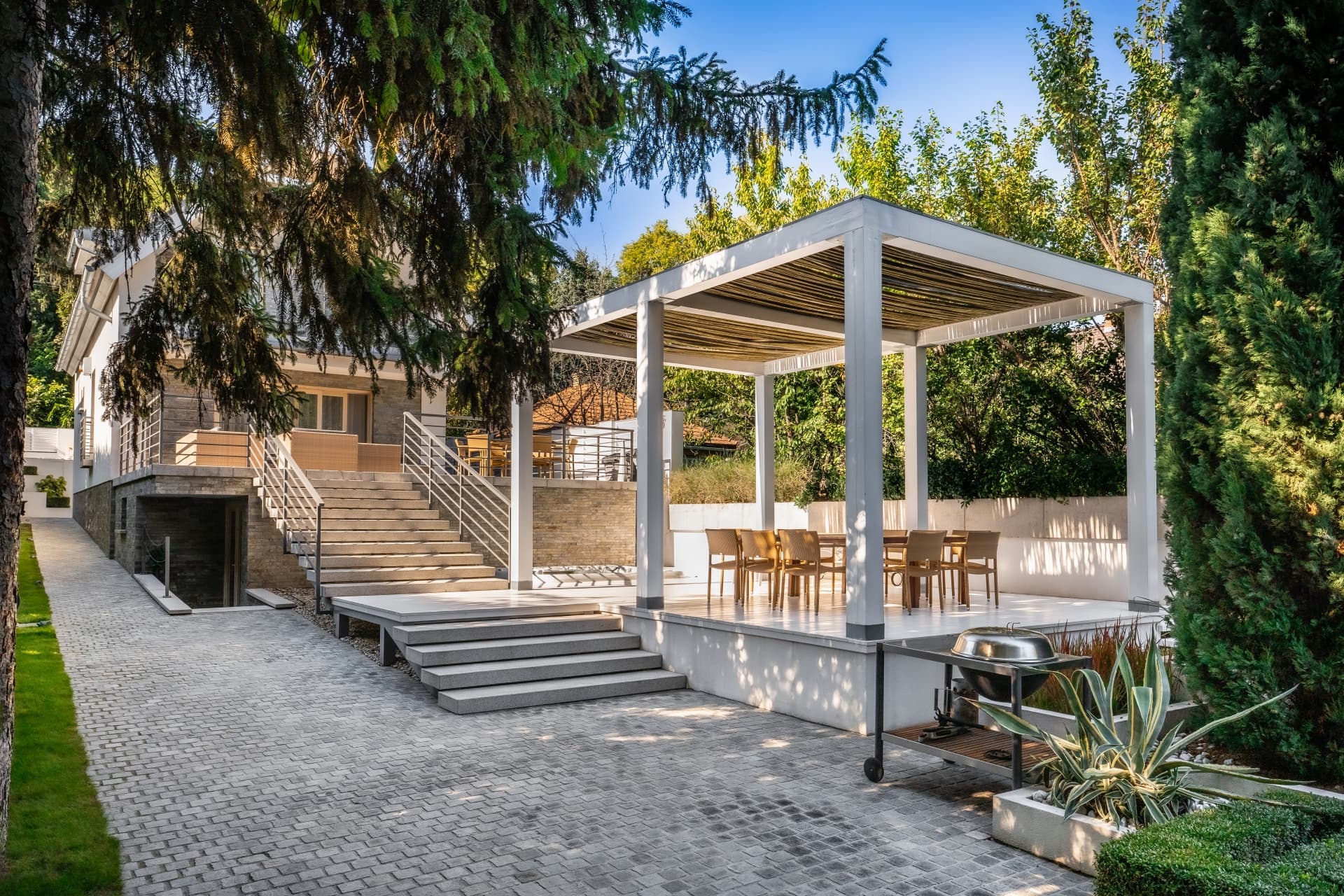 Moderner Hinterhof mit weißer Pergola, Esstisch, Steintreppe und üppigen grünen Bäumen im Sonnenlicht.