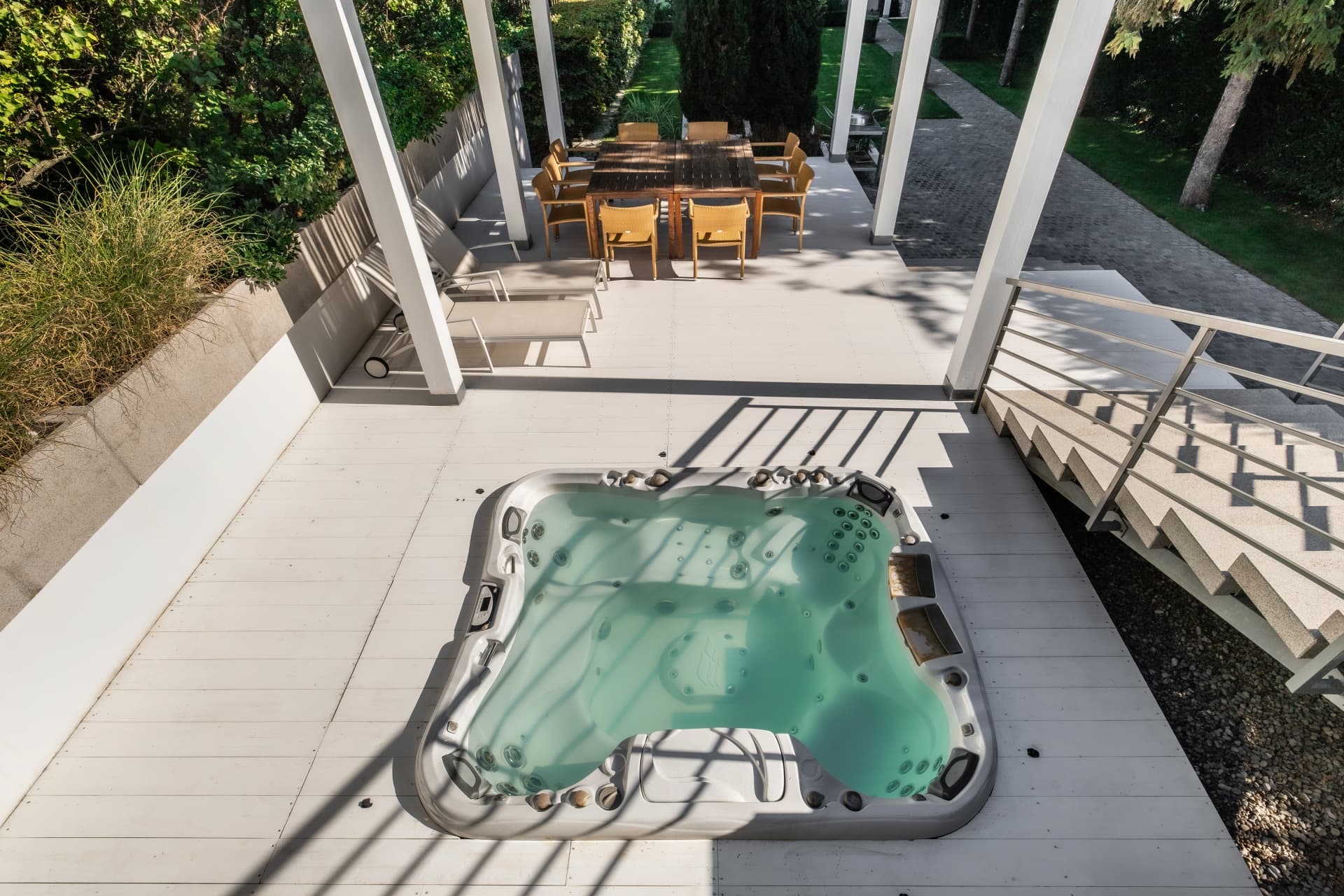 Quadratischer Whirlpool auf einer weißen Holzterrasse mit Sitzgruppe und Liegestühlen im Hintergrund.