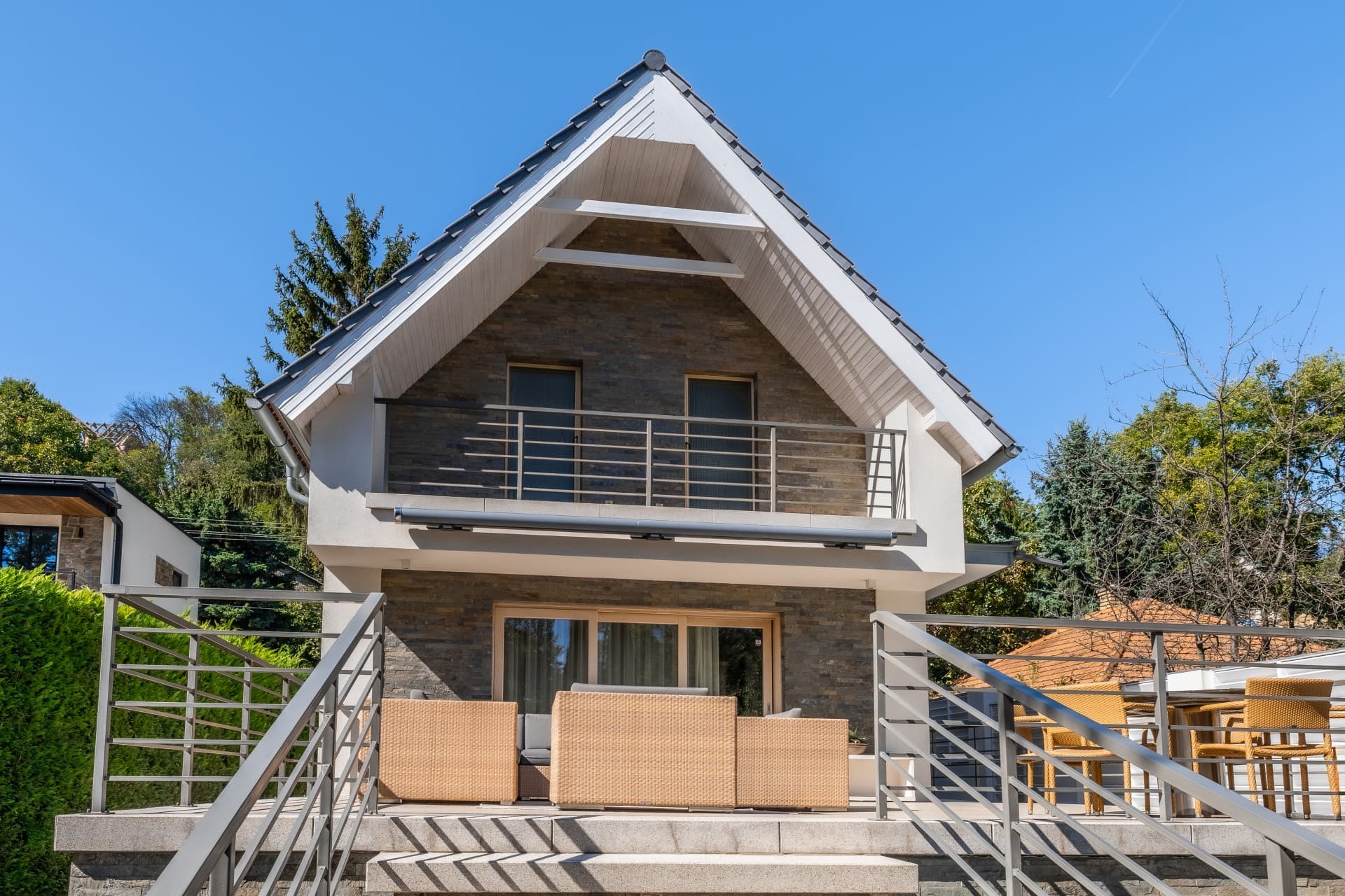 Modernes Haus mit Steinfassade, steilem Giebeldach, Balkon und einer Terrasse mit beigen Korbmöbeln.